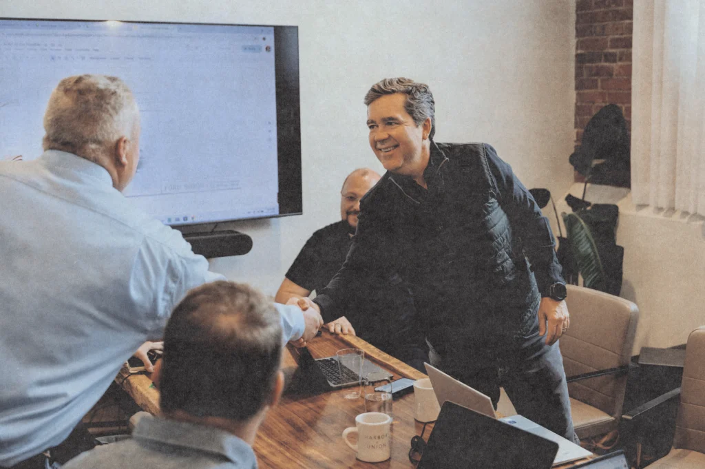 Four men are meeting in a conference room. Two of them are shaking hands and smiling, while the others sit at a table with laptops, coffee mugs, and documents. A large screen is visible in the background.