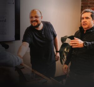 Two men greet a third man with a handshake in a conference room. A presentation is displayed on a screen behind them, showing a newspaper headline. Another man stands nearby, reaching out his hand.