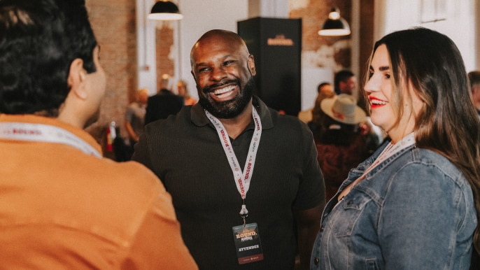 Three attendees wearing event lanyards engaged in conversation inside a warm, industrial-style venue, capturing the collaborative energy