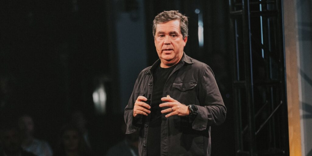 Mike Hollis standing on a dimly lit stage, using expressive hand gestures while presenting to an audience, illustrating themes of collaboration and innovation that support harmonizing perspectives around healthcare AI.