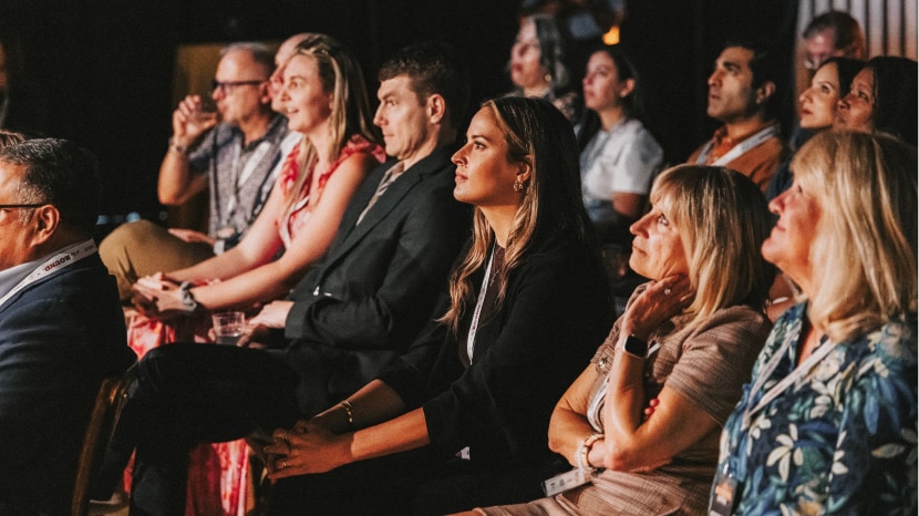 A seated audience attentively watching a presentation in a warmly lit venue, capturing a moment of focused engagement