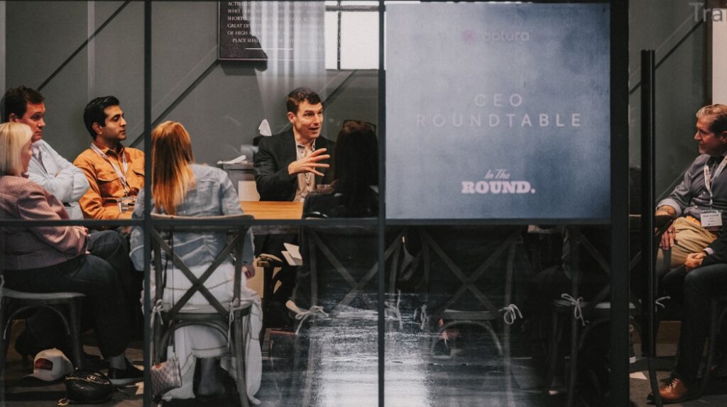 Participants seated around a table in a glass-walled room engaged in a focused CEO roundtable discussion during the In the Round Optura event, highlighting collaborative dialogue and strategic idea‑sharing