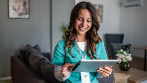 A smiling female nurse in teal scrubs looking down and interacting with a digital tablet.
